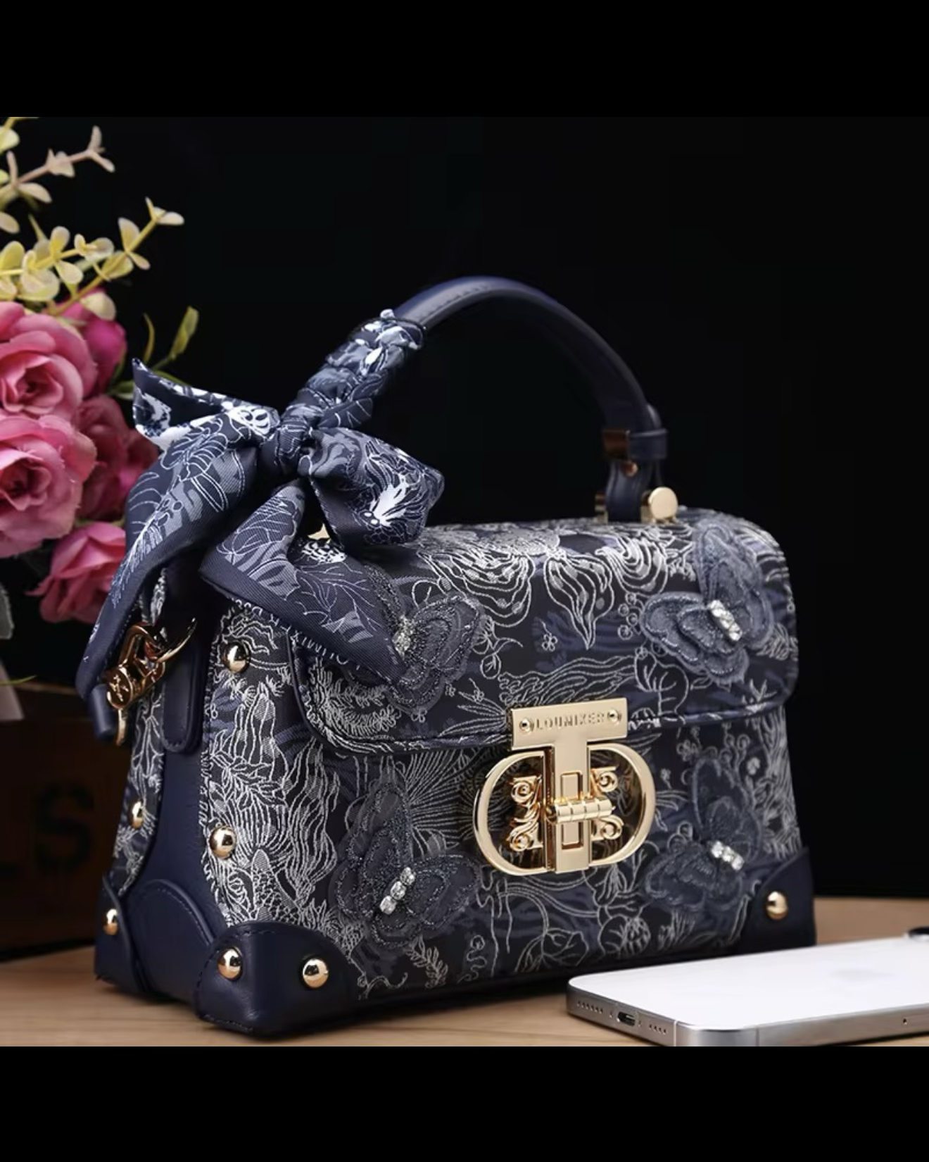 Decorative handbag with floral patterns and a bow, placed on a surface with flowers and a phone in the background.