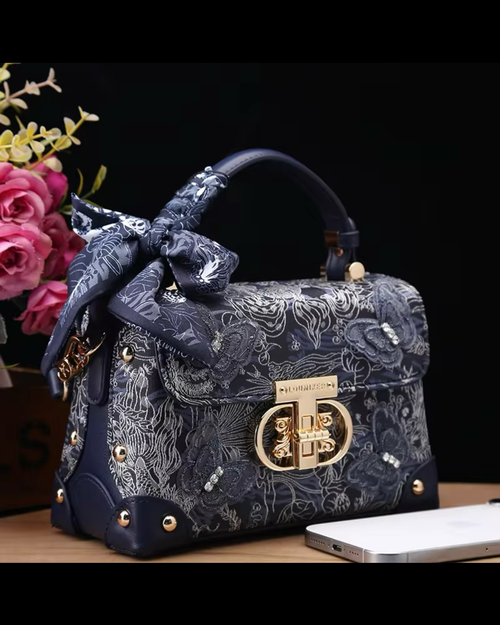 Decorative handbag with floral patterns and a bow, placed on a surface with flowers and a phone in the background.