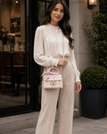Woman in a white blouse and beige pants holding a pink handbag outdoors.