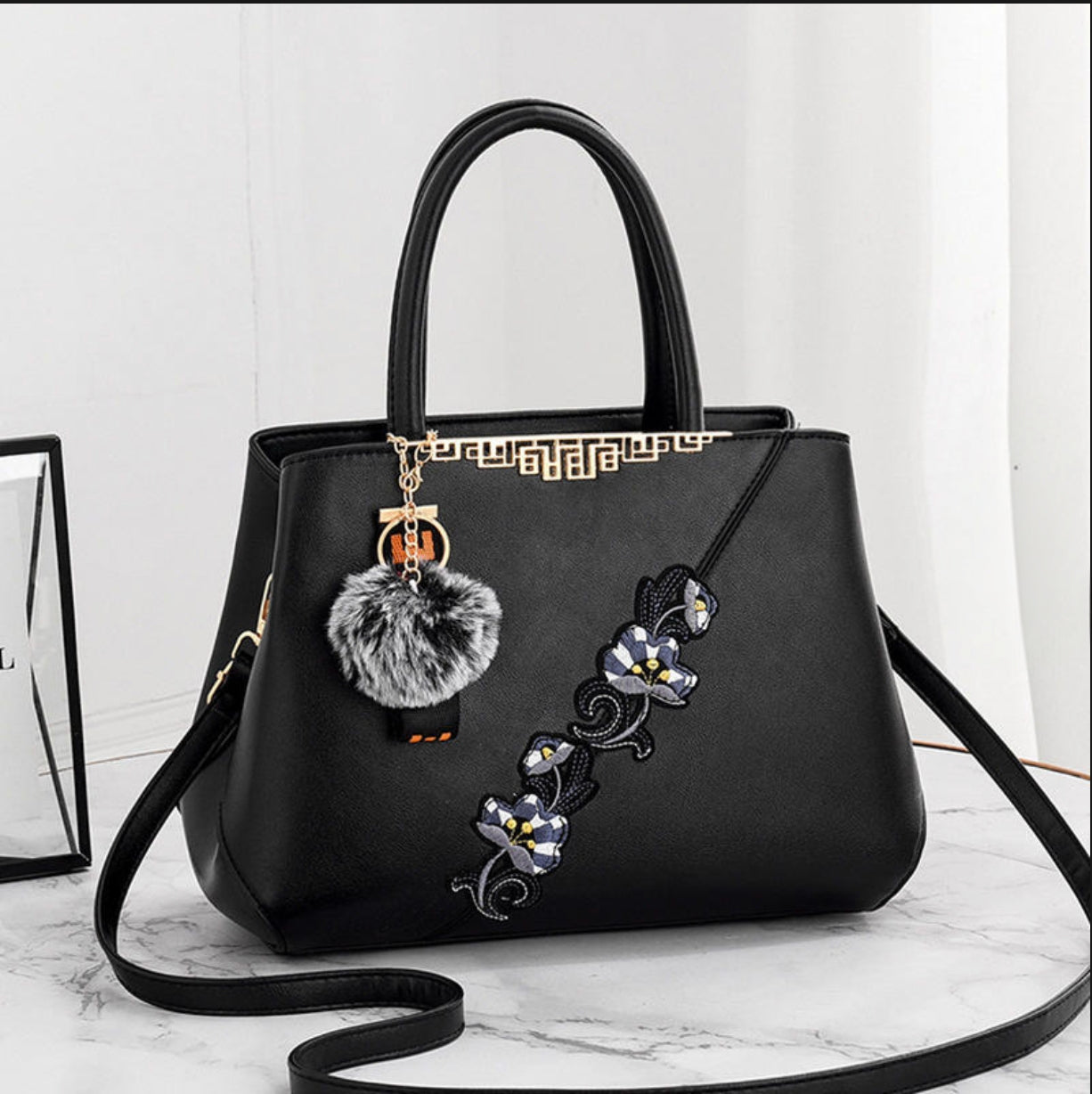 Black handbag with floral embroidery and a pom-pom charm on a white background